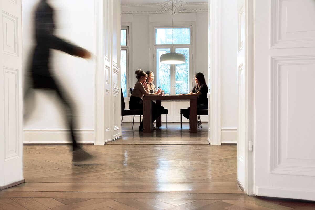 Twentyone Brands Einblick in die Agentur, eine Mitarbeiterin undscharf in Bewegung und weitere Mitarbeiterinnen bei einer Besprechung im Konferenzraum.